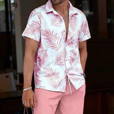 A man wearing a white short-sleeve button-up shirt with a pink palm leaf pattern and solid pink shorts, holding a brown paper shopping bag.