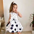 Child wearing a white dress with black polka dots, standing indoors.