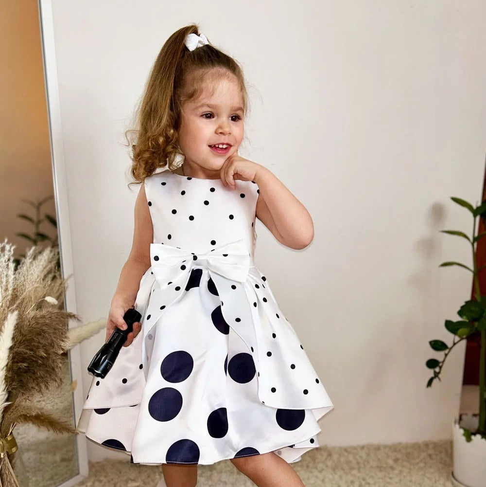 Child wearing a white dress with black polka dots, standing indoors.