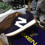 Navy blue and white sneaker held by a hand with a blurred background of shoes and packaging.
