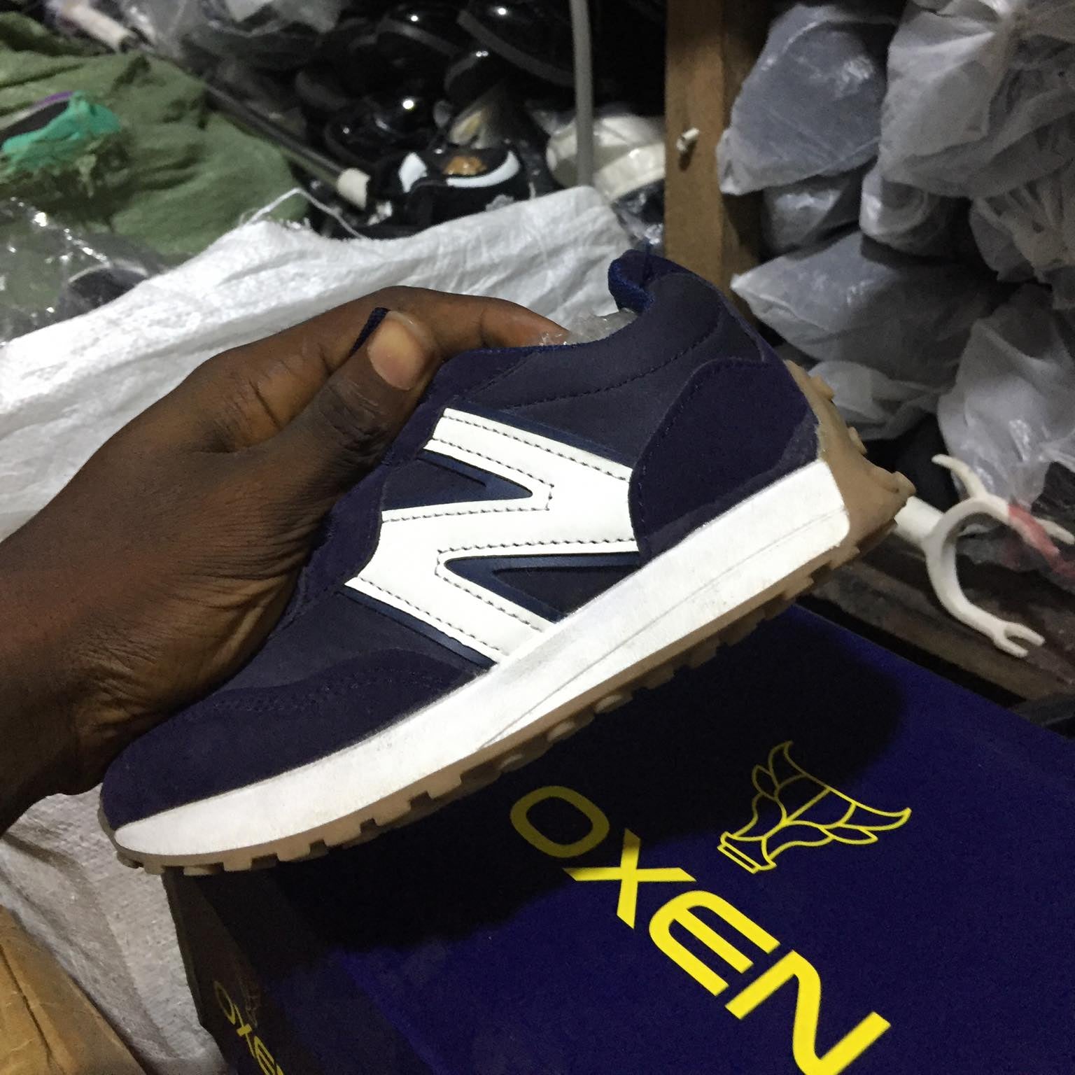Navy blue and white sneaker held by a hand with a blurred background of shoes and packaging.