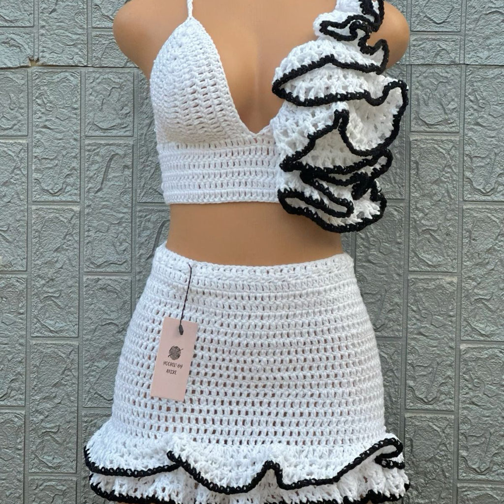 Mannequin wearing a white crochet outfit with black trim against a textured gray wall.