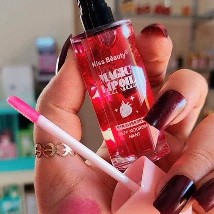 Hand holding a red lip oil with a brush, labeled 'Kiss Beauty Magic Lip Oil', against a blurred background.