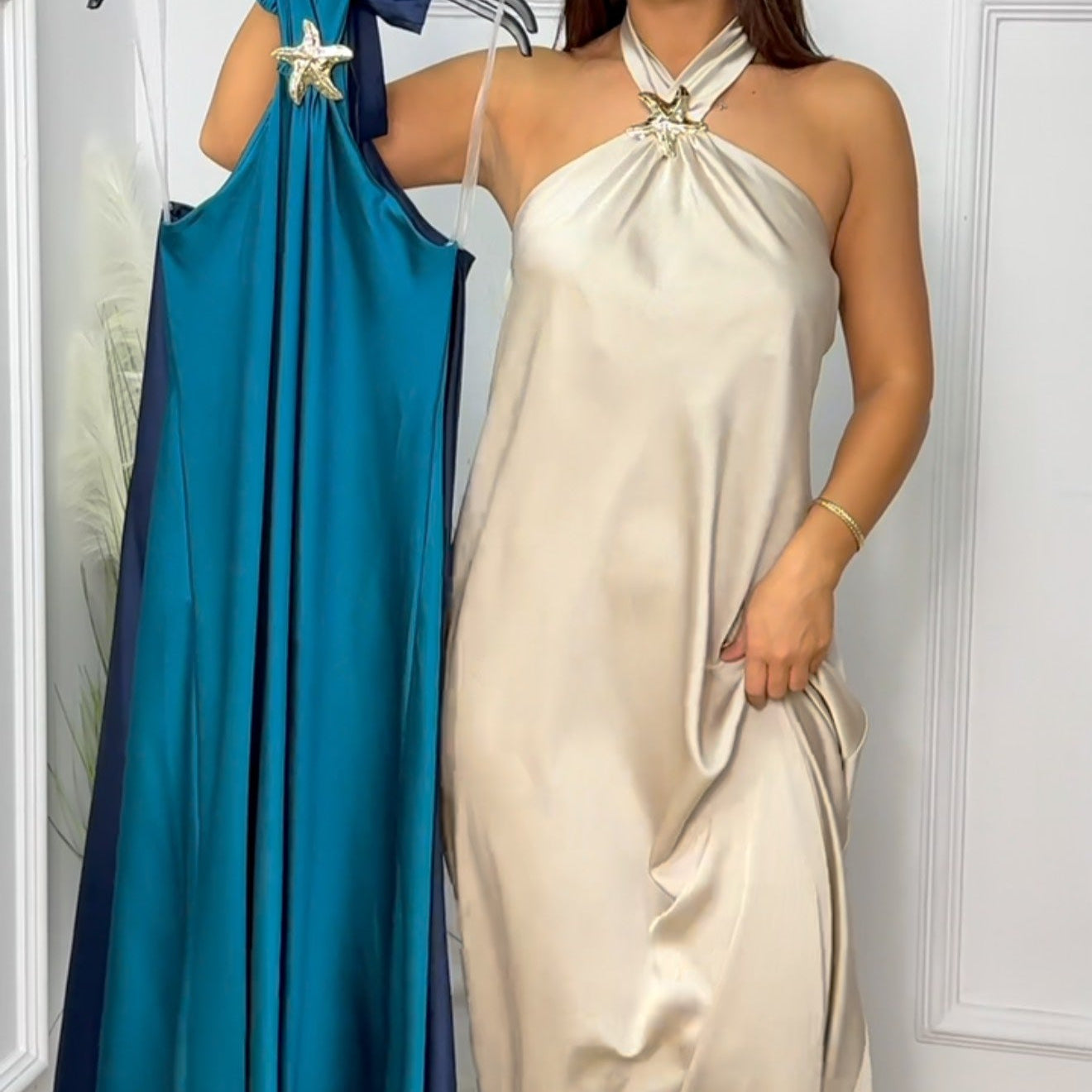 Woman holding a beige dress and a blue dress against a white wall.