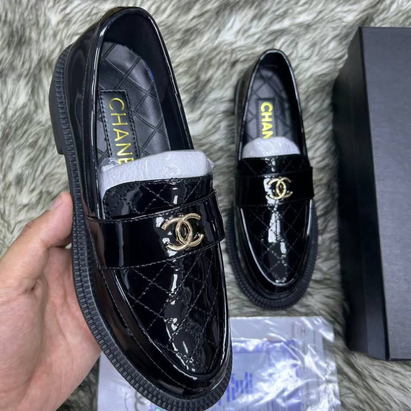 Black patent leather Chanel loafers on a textured surface with packaging.