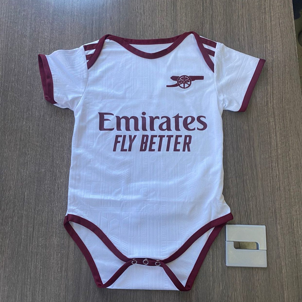 White baby onesie with red accents and 'Emirates Fly Better' text on a wooden surface.