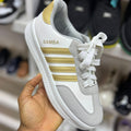 White and gold sneaker with 'Samba' branding held in a hand against a blurred shoe store background.