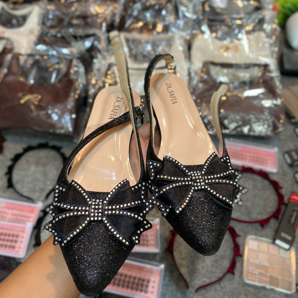 Black glittery high-heeled shoes with bow details held in front of a display of handbags.