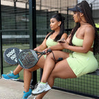 Two women sitting on a bench at a padel court, wearing matching light green athletic sets and holding padel rackets.