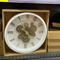 Decorative clock with gear mechanism in a beige box on a shelf.