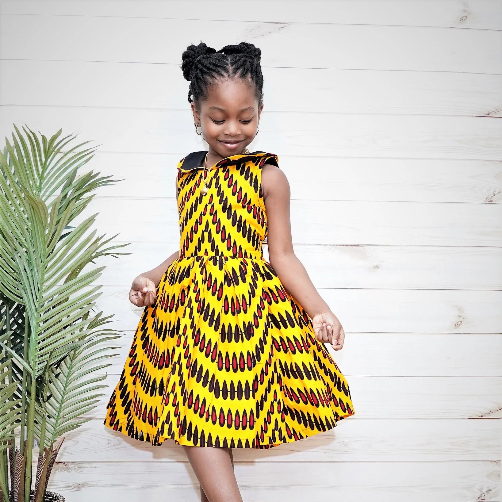 Young girl wearing a yellow and black patterned dress standing indoors with a plant in the background.