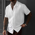 A man wearing a white short-sleeve button-up shirt with a textured knit pattern, posed against a dark gray wall.
