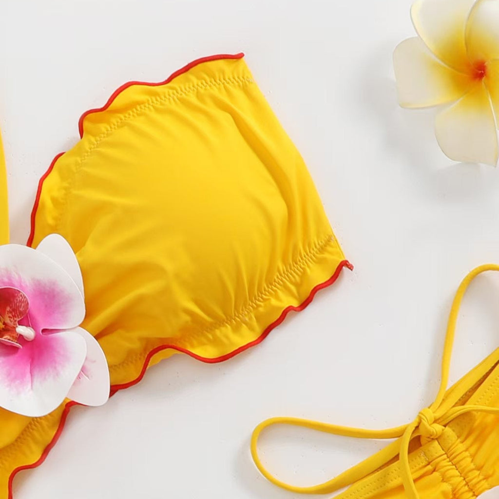 Yellow bikini top and matching bottoms laid flat on a white background, styled with pink-and-white flowers. The bikini top has a ruffled edge with a thin red trim.