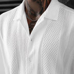 A close-up of a man wearing a white textured knit button-up shirt with a collar and a silver chain necklace.