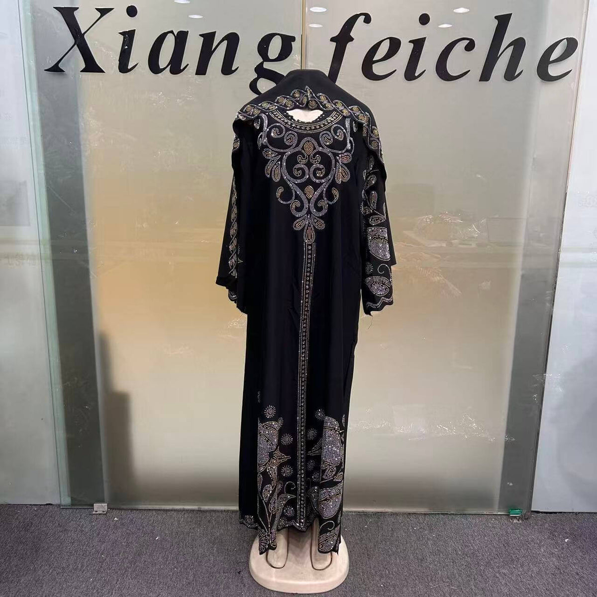 A long black abaya dress with ornate silver and gold embroidery, displayed on a mannequin in front of a glass door with the text 'Xiang feiche' visible.