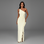 A woman modeling a floor-length, one-shoulder cream gown with ruching and layered ruffle details on the skirt, photographed against a seamless gray studio background.