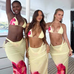 Three women posing on a yacht deck wearing matching pale yellow bikini sets with vibrant pink and red floral prints on the sarongs.