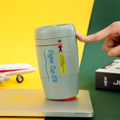 A light gray engine cup with blue and red printed graphics sits on top of a silver laptop. A human hand is touching the cup, and the background includes a bright yellow wall, a green surface, a toy airplane, and a box.