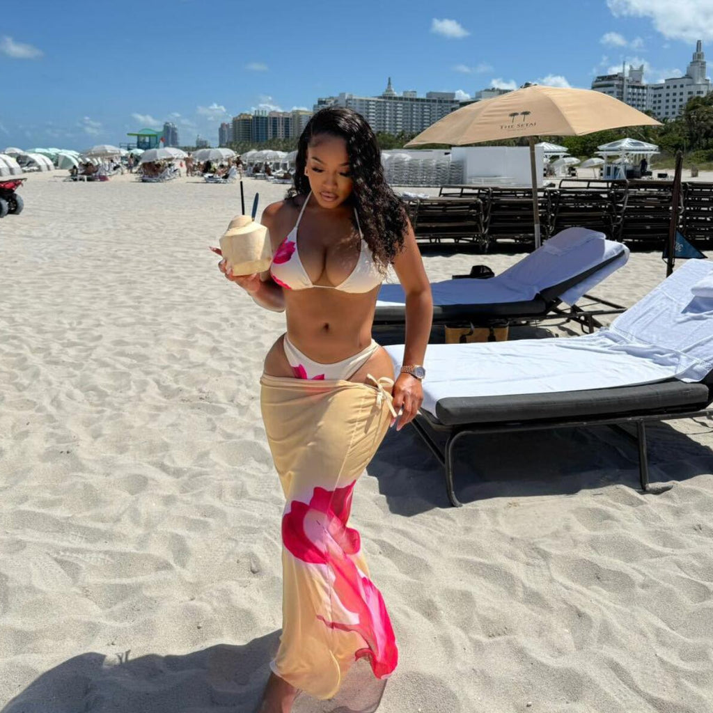A woman walks on a sandy beach wearing a white bikini with pink floral details and a matching beige sarong with bright pink accents. She is holding a coconut drink, with beach chairs, umbrellas, and buildings in the background under a blue sky.