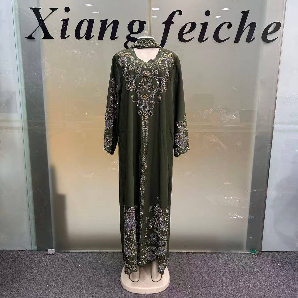 An olive green long kaftan dress with intricate silver and gold embroidery, displayed on a mannequin in front of a glass door with 'Xiang feiche' written on it.