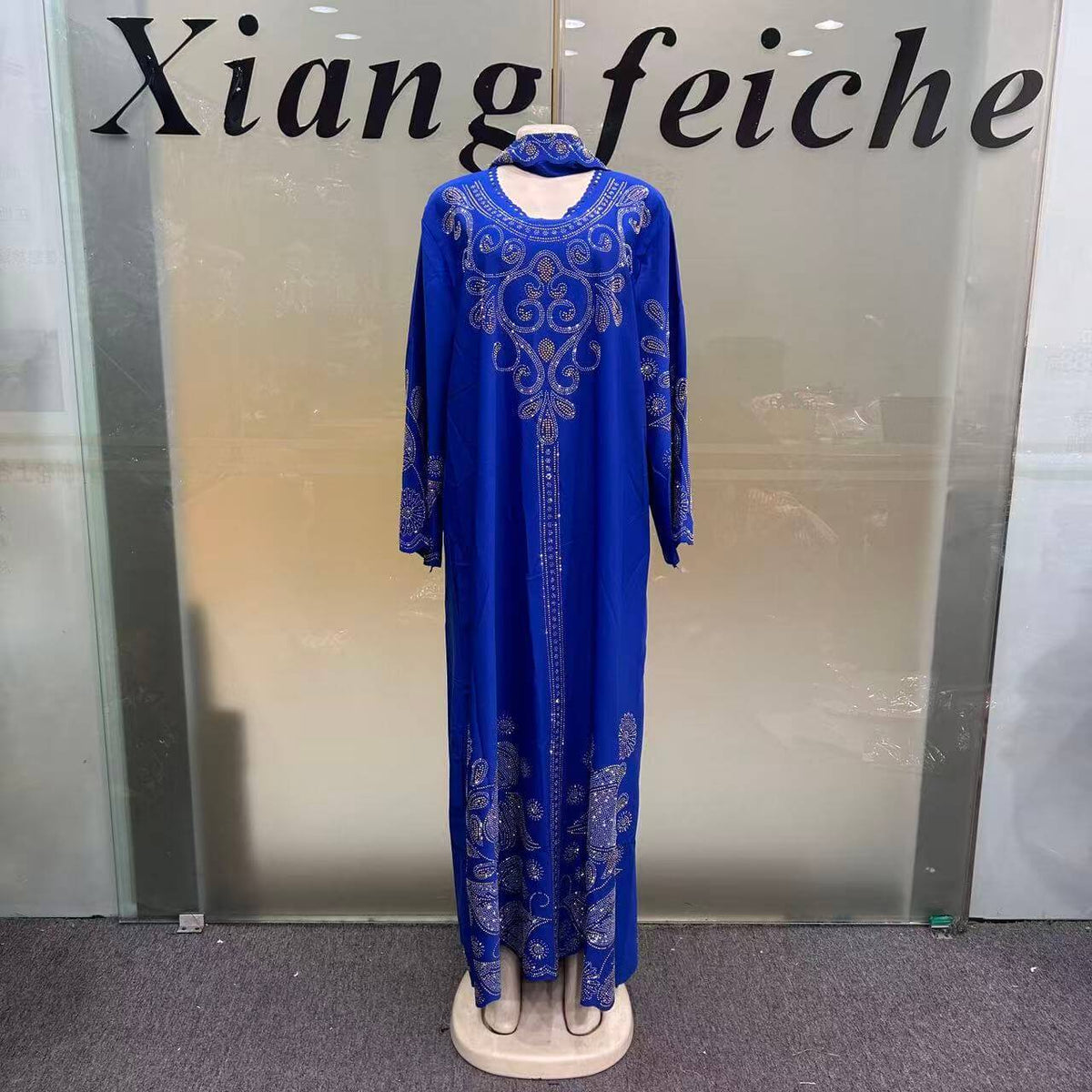 A royal blue long caftan dress with intricate silver embroidery, displayed on a mannequin in front of a glass storefront window with the text 'Xiang feiche' visible.