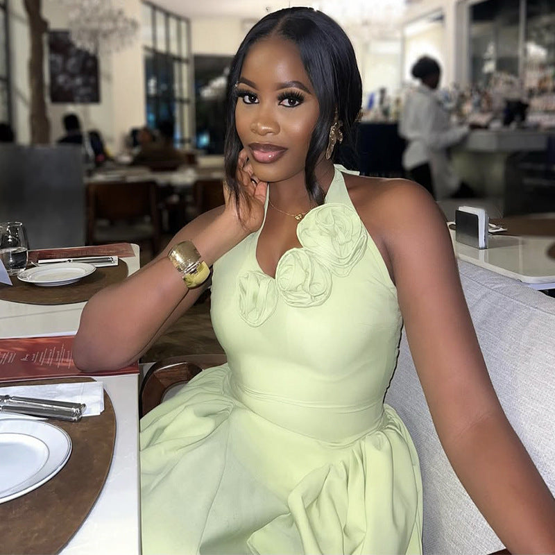 A woman posing in a restaurant while wearing a light sage green halter dress with large fabric flowers on the bodice.