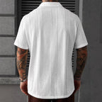 A man viewed from the back wearing a white short-sleeve button-up shirt with vertical textured knit panels, standing against a gray wall.