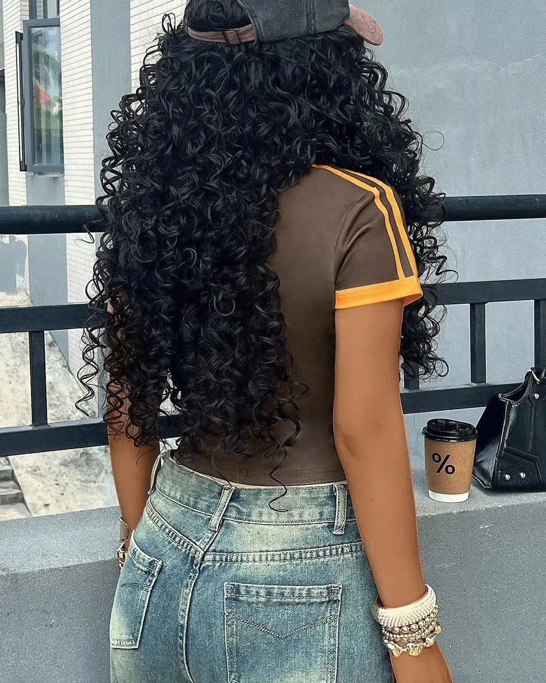 Person with long curly hair wearing a cap and casual outfit, standing on a balcony.