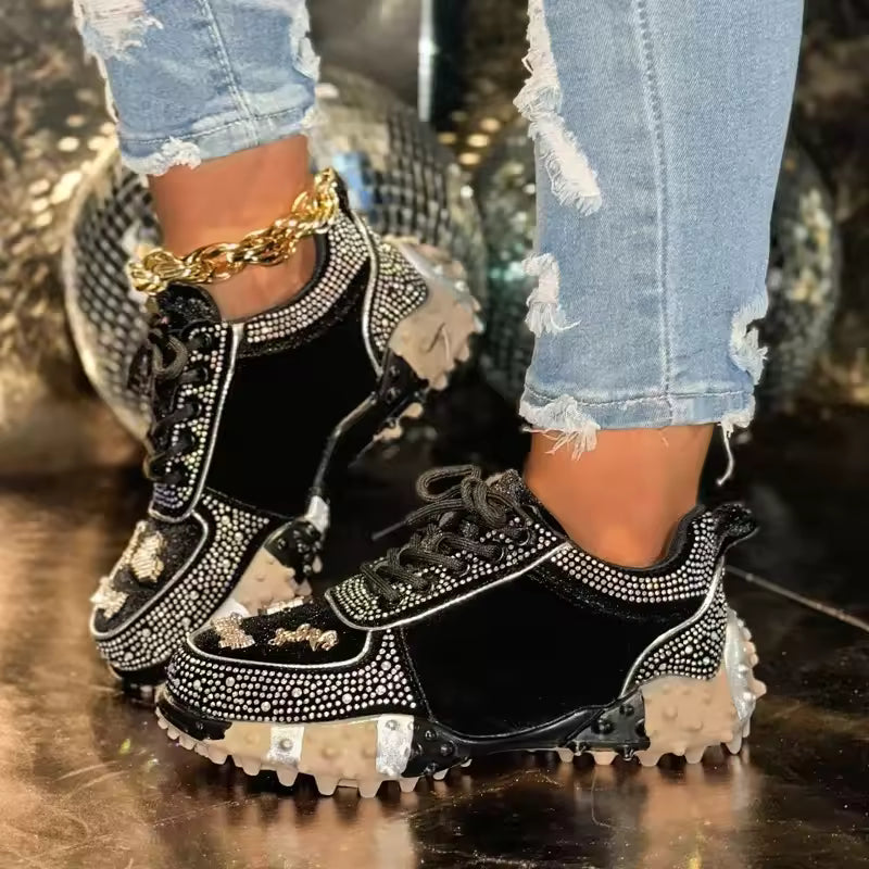 Black sneakers with rhinestone details worn with ripped jeans on a reflective surface.
