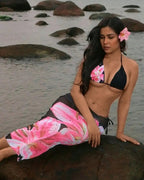 Woman in a black bikini with pink floral accents sitting on rocks by the water.