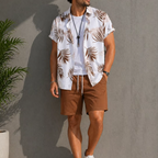 A man wearing a white short-sleeve button-up shirt with a brown palm leaf print, a white t-shirt underneath, and brown drawstring shorts, standing against a gray wall.
