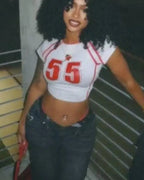 Woman wearing a white crop top with red numbers and dark jeans, standing indoors.