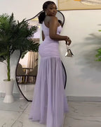 Woman in a long purple dress holding a handbag indoors with a plant and mirror in the background.