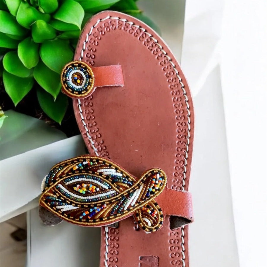 Brown sandal with decorative strap on a white surface with green plant in the background