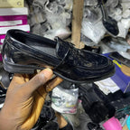 Black shiny loafer held by a hand with a cluttered background