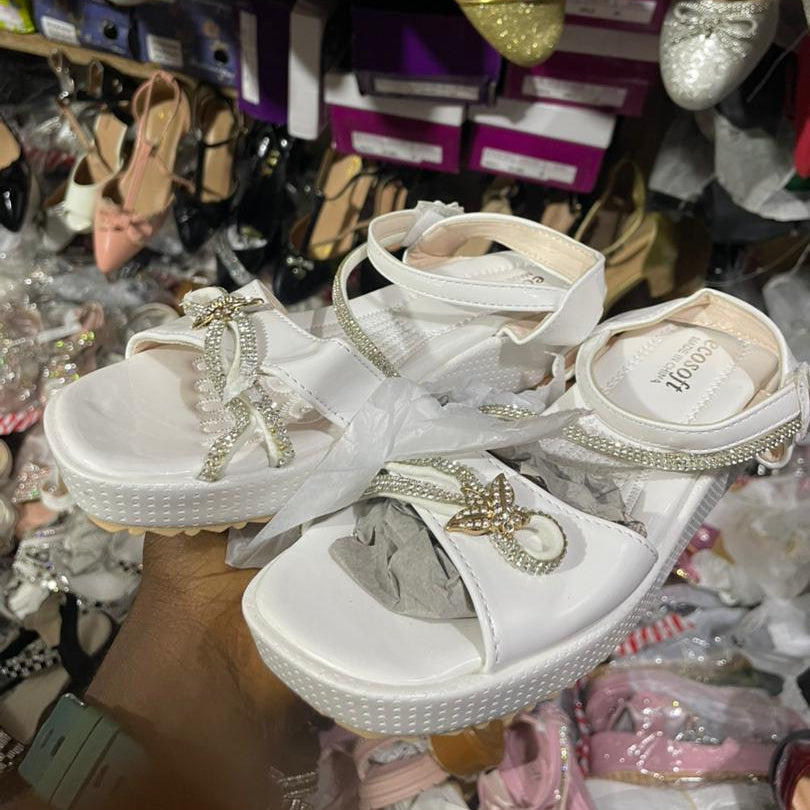 White sandals with decorative elements held in front of a display of various shoes.