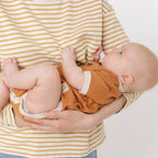 Person holding a baby wearing a striped shirt and orange outfit against a plain background
