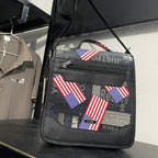 A black crossbody bag with American flag patches and collage-style text graphics, held by a person's hand on a store display shelf.