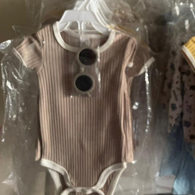 Two baby rompers on hangers with tags, one brown and one yellow, in a clear plastic bag.