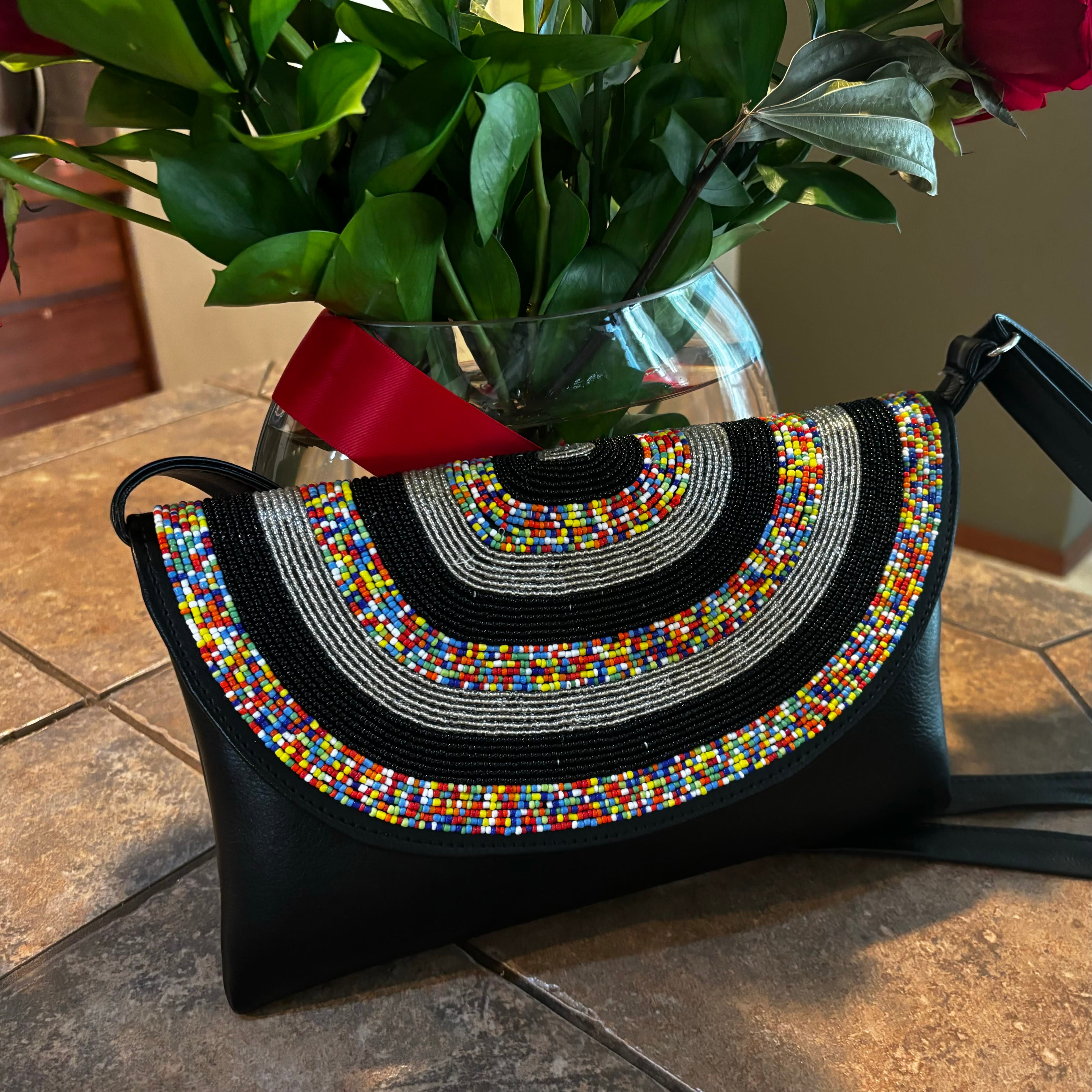 Black handbag with colorful beaded design on a tiled floor with a vase of flowers in the background.