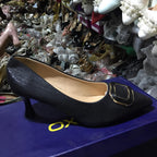 Black high-heeled shoe with gold decorative element on a blue surface, surrounded by other shoes.