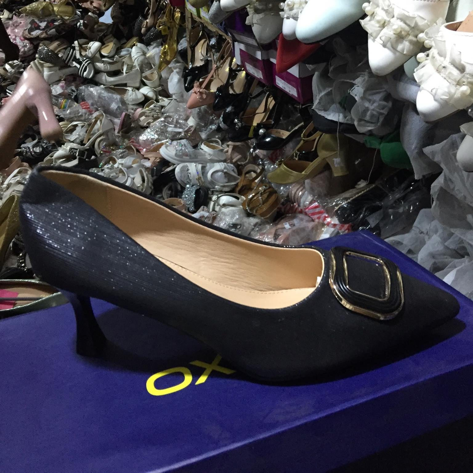 Black high-heeled shoe with gold decorative element on a blue surface, surrounded by other shoes.
