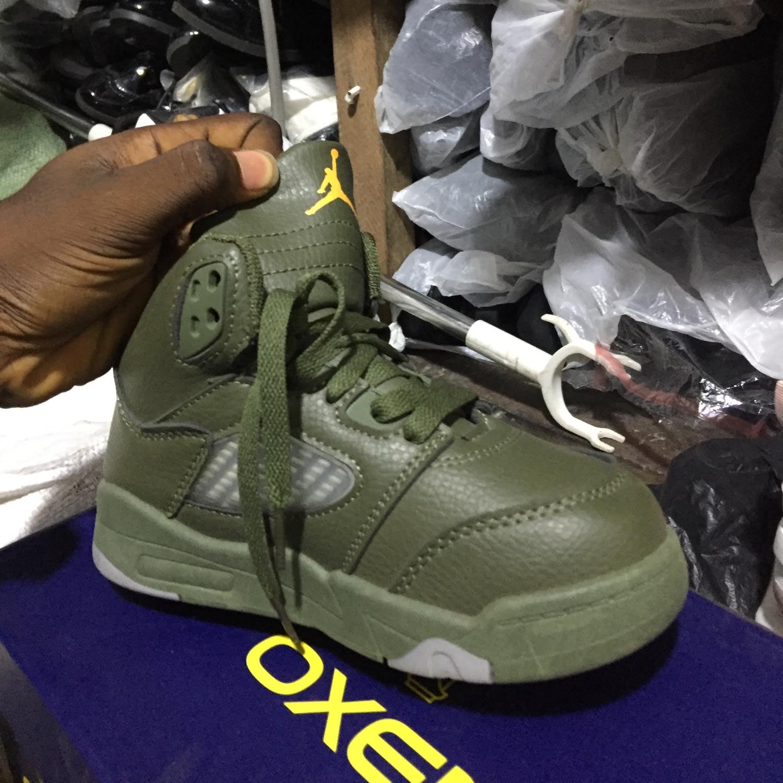 Green sneaker held by a hand with a blurred background of shoes and plastic bags.