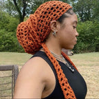 Woman wearing an orange knitted headscarf and matching scarf outdoors with trees in the background