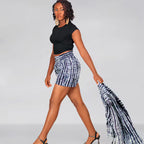 Woman wearing a black top and patterned shorts holding a matching scarf against a gray background