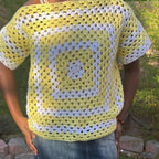 Person wearing a yellow and white crocheted square pattern top outdoors.