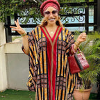 Woman in a patterned dress and red headscarf standing outdoors with plants and a building in the background.