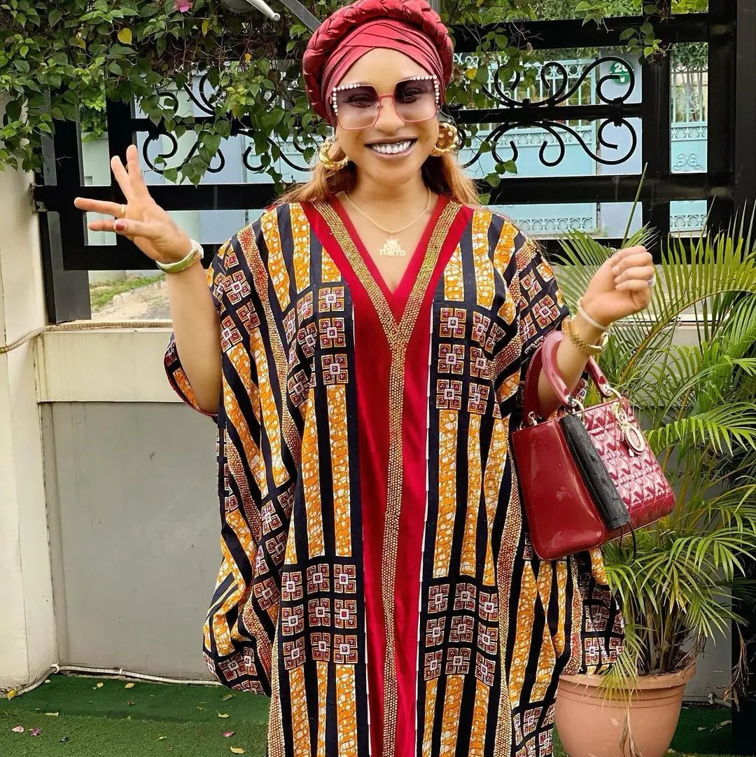 Woman in a patterned dress and red headscarf standing outdoors with plants and a building in the background.